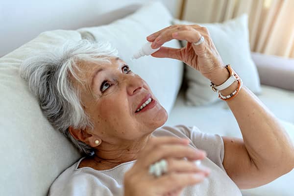 Old woman applying eye drops on her eyes after Cataract Surgery while laying in bed