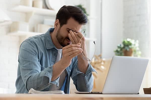 Man rubbing his eyes in front of a laptop due to dry eye condition